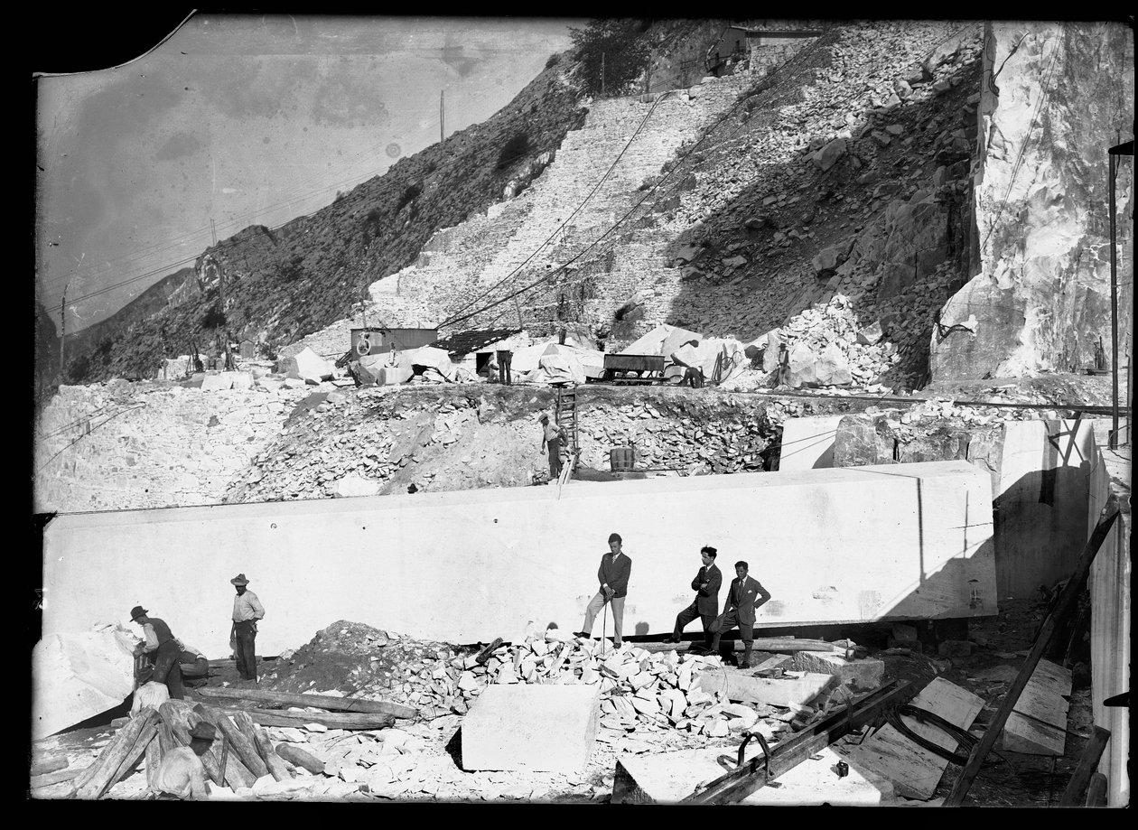Trois hommes observent le travail de plusieurs excavatrices dans les Alpes apuanes. Derrière eux se trouve un énorme bloc de marbre déjà taillé et prêt à être transporté dans la vallée - A. Corsini