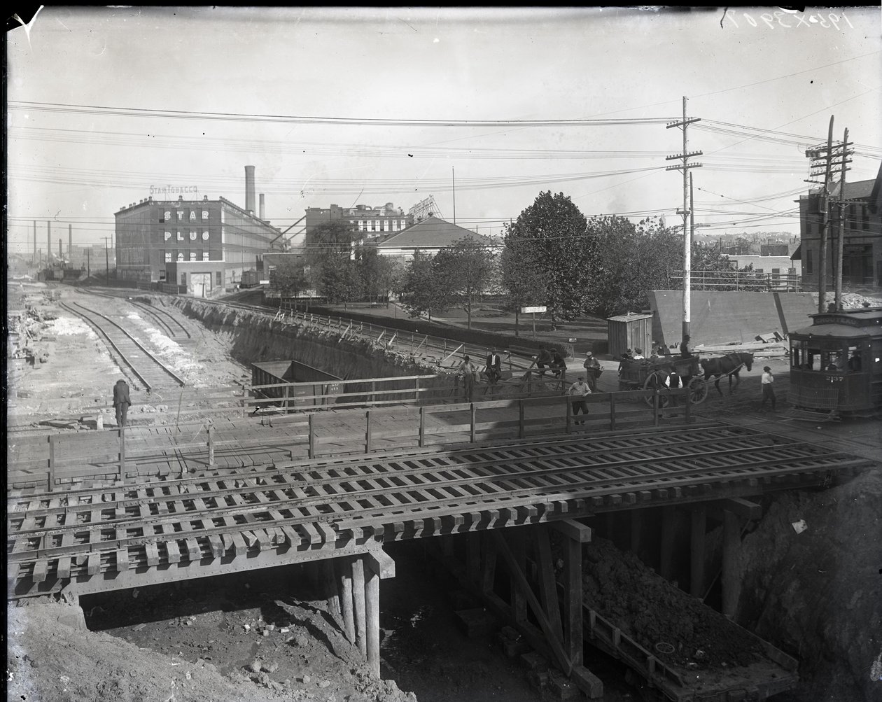 Photographie horizontale en noir et blanc montrant un pont en bois et des voies de tramway sur des voies ferrées partiellement construites. Un tramway marqué Chouteau 2137 commence à traverser le pont à l