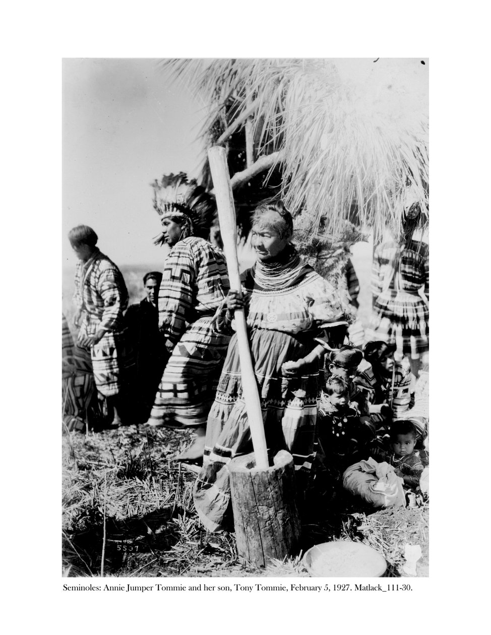 Annie Jumper Tommie utilisant un mortier et un pilon pour faire de la farine de maïs, et son fils, Tony Tommie, portant un bonnet de plumes d
