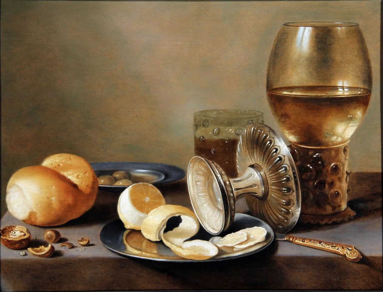 Banketje monochrome avec deux plats en étain, une tazza en argent renversée, un couteau en vermeil, un verre à bière, un verre à vin, un petit pain, des noix, des olives et un demi-citron pelé. - Hans van Sant