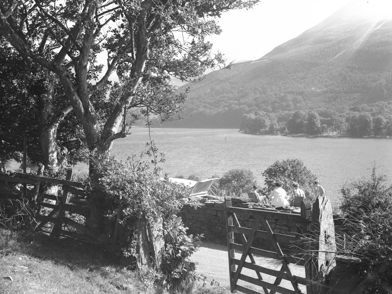 Quatre personnes assises sur un muret de pierres sèches en train de pique-niquer avec vue sur le lac, voiture garée avec coffre ouvert. - Joseph Hardman