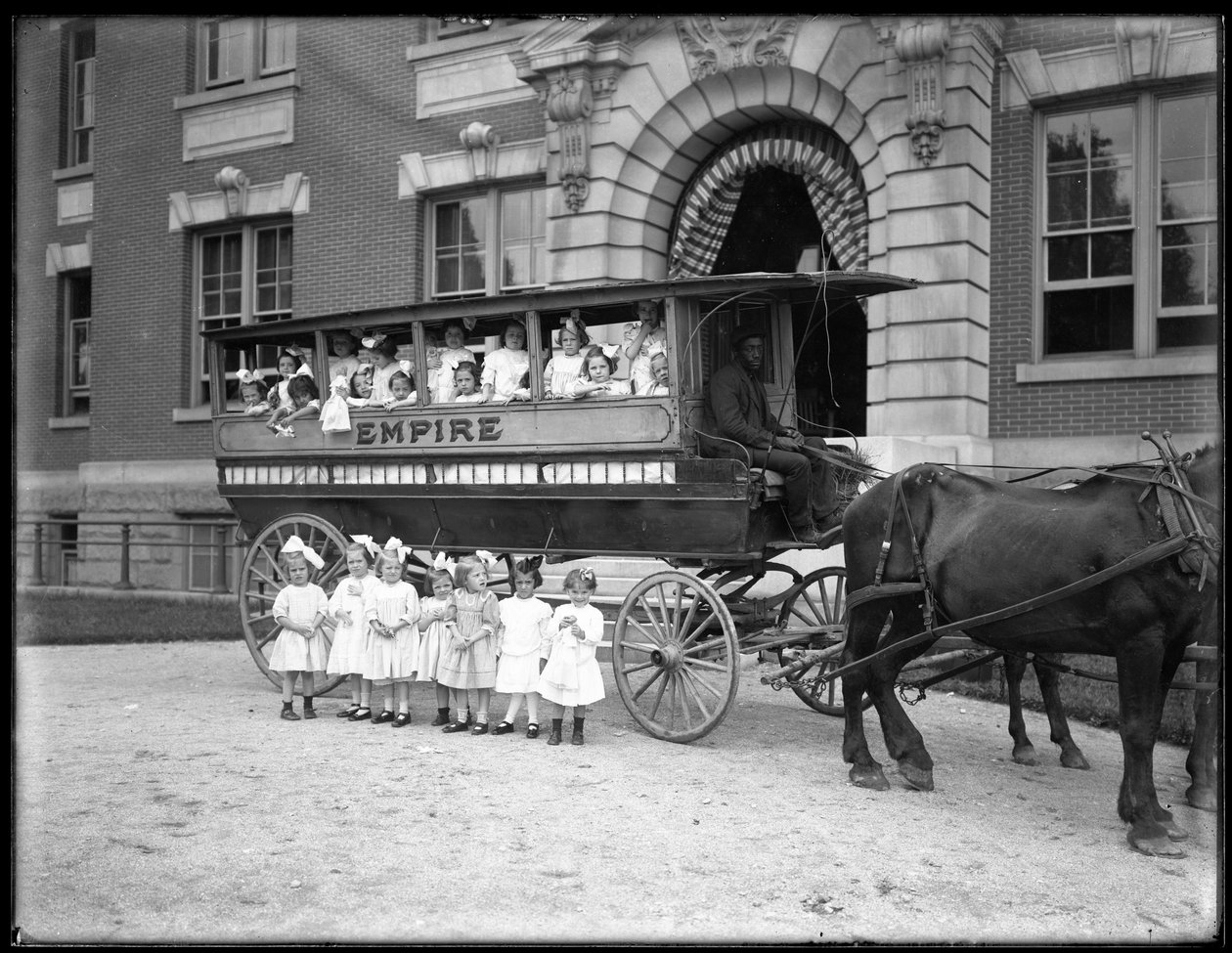 Petites filles posant dans et devant une charrette tirée par des chevaux, devant un bâtiment non identifié, probablement l