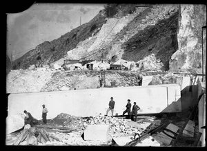 Trois hommes observent le travail de plusieurs excavatrices dans les Alpes apuanes. Derrière eux se trouve un énorme bloc de marbre déjà taillé et prêt à être transporté dans la vallée