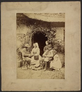 [Groupe assis devant la porte ouverte : deux hommes en chapeau, une femme en bonnet, deux jeunes filles - l