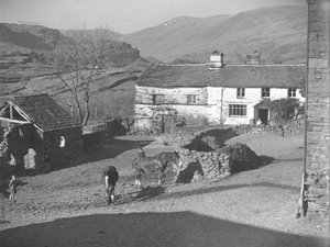 Trois jeunes garçons surveillant des chevaux dans la cour de ferme de Kentmere, avec la ferme et les montagnes derrière.