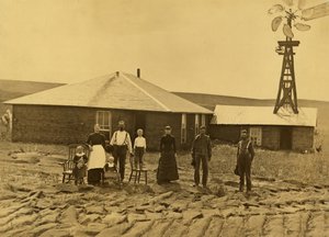 Famille de colons devant leur maison en terre avec un moulin à vent sur le toit du bâtiment adjacent dans le comté de Custer, Nebraska, vers 1885 (photo n/b) - Solomon D. Butcher