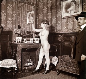 Photographie énigmatique montrant une jeune femme complètement nue (sauf ses chaussures et ses bas) recoiffant ses cheveux devant un miroir en présence d