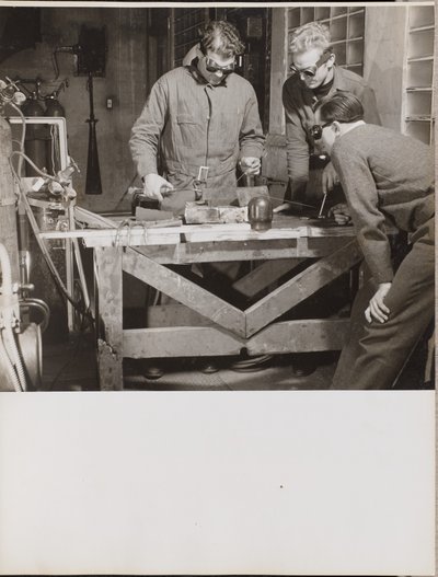 [Trois hommes portant des lunettes de protection soudent une pièce de métal sur une table]. - Arnold Eagle