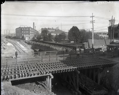 Photographie horizontale en noir et blanc montrant un pont en bois et des voies de tramway sur des voies ferrées partiellement construites. Un tramway marqué Chouteau 2137 commence à traverser le pont à l