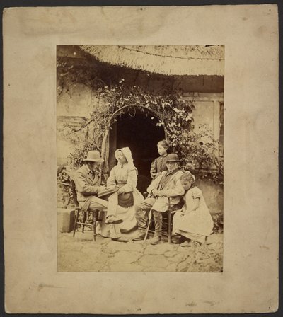 [Groupe assis devant la porte ouverte : deux hommes en chapeau, une femme en bonnet, deux jeunes filles - l
