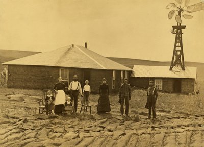 Famille de colons devant leur maison en terre avec un moulin à vent sur le toit du bâtiment adjacent dans le comté de Custer, Nebraska, vers 1885 (photo n/b) - Solomon D. Butcher