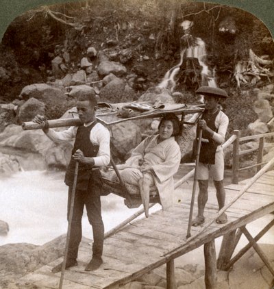 Femme japonaise dans une chaise de montagne yamakago traversant la rivière torrentielle Daiya près de Nikko, Japon - Underwood and Underwood