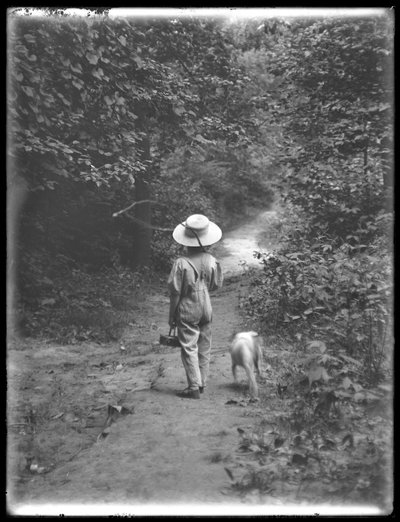 William Gray Hassler en chapeau de paille et salopette, portant une canne à pêche et un panier, marchant sur un chemin dans les bois avec un petit chien (Bessie), 20 août 1911 - William Davis Hassler