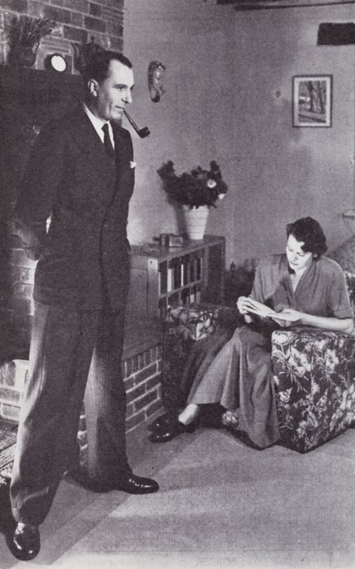 La femme lit, tandis que le mari se réchauffe le dos contre le feu, vers 1950 - English Photographer