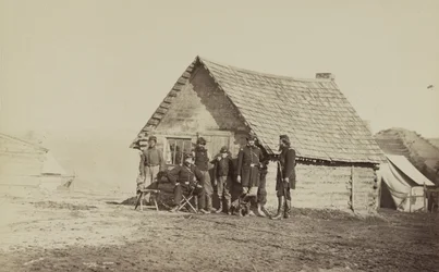 Un groupe de soldats et deux jeunes hommes, dont un afro-américain, devant des quartiers en cabane en rondins, 1861-65