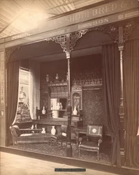 Exposition de James Shoolbred & Co. -- Bâtiment Principal, scène de salon comprenant un buffet et un miroir avec des vases dessus et devant, des chaises, une table et un canapé. Sur le sol, un tapis et les murs sont couverts de papier peint floral