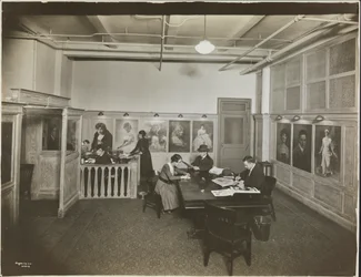 Bureau principal/salle de réception à la Mutual Film Co. avec secrétaire au bureau, personnes à une table lisant, et portraits sur le mur, New York