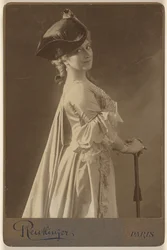 [Femme non identifiée portant un chapeau noir, debout, les mains posées sur une canne].