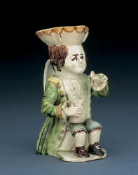 Pichet Toby du buveur, son chapeau au bord festonné brun et jaune, ses cheveux bruns incisés noués en queue avec une rosette blanche, assis avec une cruche en balustre sur le genou, vers 1770