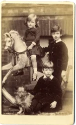 Jeune fille sur un cheval à bascule avec un garçon debout et un garçon assis avec un chien