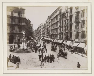 Vue de la Via Roma à Naples avec une statue, des magasins, des voitures à cheval et des passants