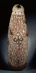 Bouclier Asmat ovale, sculpté en relief avec des ornements en volutes incorporant des figures hautement stylisées, franges tressées à la bordure