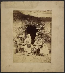 [Groupe assis devant la porte ouverte : deux hommes en chapeau, une femme en bonnet, deux jeunes filles - l