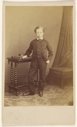 [Petit garçon non identifié en costume, debout devant une table sur laquelle sont posés des livres].