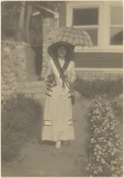 [Femme debout dans le jardin devant la maison, avec un appareil photo et un parapluie à carreaux]