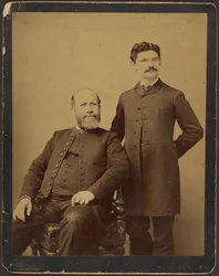 [Homme barbu non identifié assis avec homme non identifié moustachu debout]