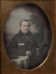 Homme portant un manteau et des gants, tenant un chapeau, assis devant un décor extérieur peint, années 1840
