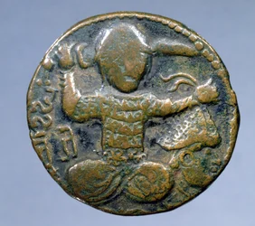 Avers de la pièce (bronze) représentant un Turc casqué assis en tailleur tenant une épée derrière sa tête dans la main droite et une tête casquée coupée dans la main gauche. frappée par Husam al-Din Yuluq Arslan (1184-1201)