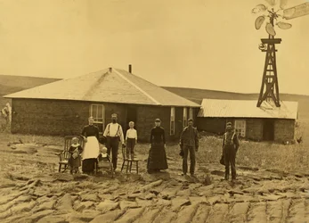 Famille de colons devant sa maison en mottes de terre, avec un moulin à vent sur le toit du bâtiment adjacent, dans le comté de Custer, Nebraska, vers 1885