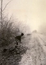 Une route de campagne enveloppée de brumes matinales. Au premier plan, un homme fend des pierres