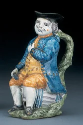 Cruche Toby avec visage et mains en manganèse pâle, tenant une cruche globulaire vide de couleur buff dans une veste et un pantalon bleu lavé, et un gilet vert, vers 1800