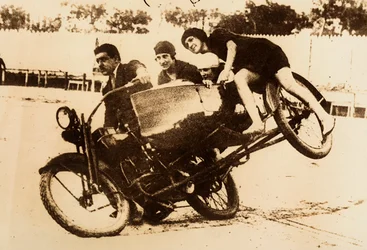 Un homme fait le malin en conduisant une moto avec un side-car transportant trois jeunes femmes. Le side-car se soulève du sol en prenant un virage.