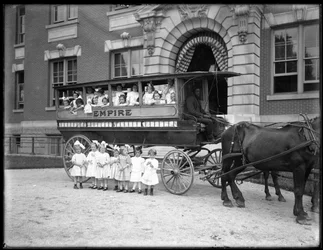 Petites filles posant dans et devant une charrette tirée par des chevaux, devant un bâtiment non identifié, probablement l
