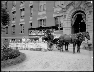 Petites filles posant dans et devant un chariot tiré par des chevaux, devant un bâtiment non identifié, probablement l