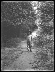 William Gray Hassler en chapeau de paille et salopette, portant une canne à pêche et un panier, marchant sur un chemin dans les bois