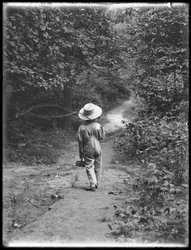 William Gray Hassler en chapeau de paille et salopette, portant une canne à pêche et un panier, marchant seul sur un chemin dans les bois, 20 août 1911